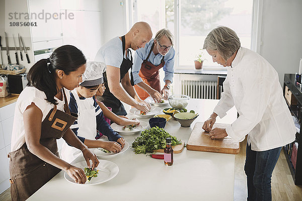 Senior woman in chef's jacket guiding family in preparing Asian food at table in kitchen