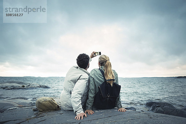 Couple taking selfie while sitting on rock by sea