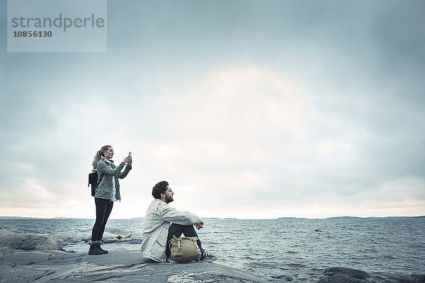 Side view of woman photographing while man sitting on rock by sea against cloudy sky