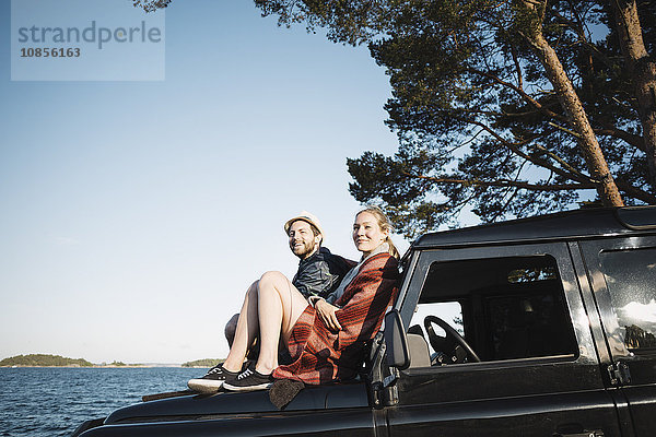 Couple sitting on jeep at lakeshore against clear blue sky