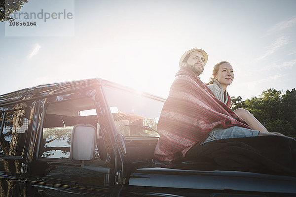 Side view of wonderlust couple wrapped in blanket sitting on jeep against sky