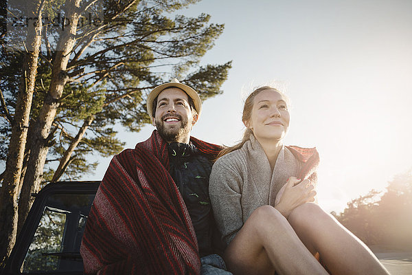 Happy wonderlust couple wrapped in blanket sitting on jeep against clear sky