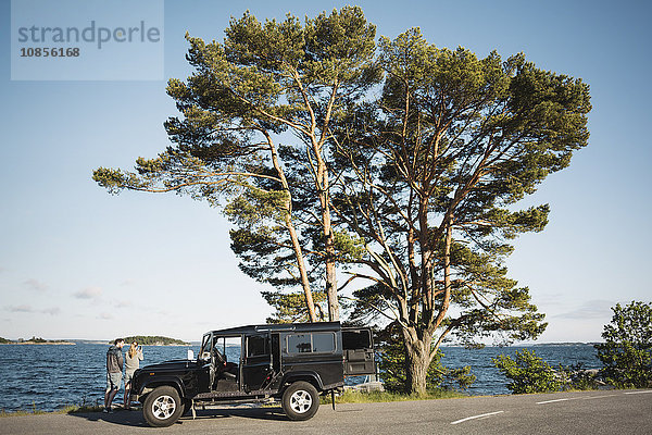 Couple standing by jeep at lakeshore against blue sky