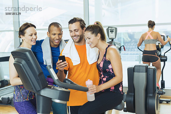 Lächelnde Freunde mit dem Handy am Heimtrainer im Fitnessstudio