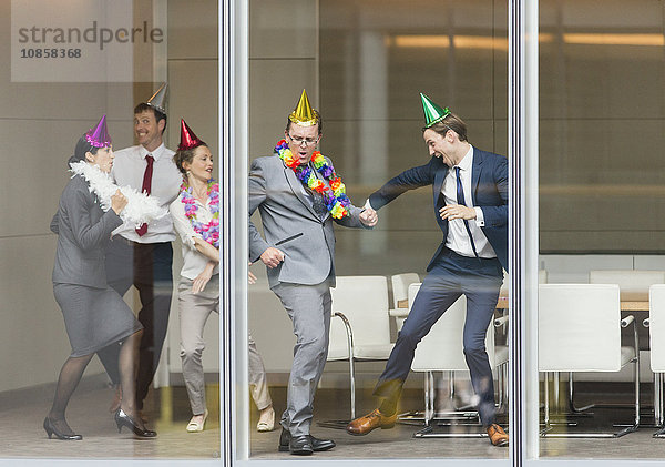 Verspielte Geschäftsleute in Partyhüten tanzen am Konferenzraumfenster
