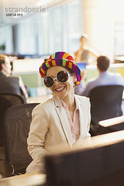 Porträt einer lächelnden Geschäftsfrau mit dummer Sonnenbrille und gestreiftem Hut  die im Büro arbeitet.