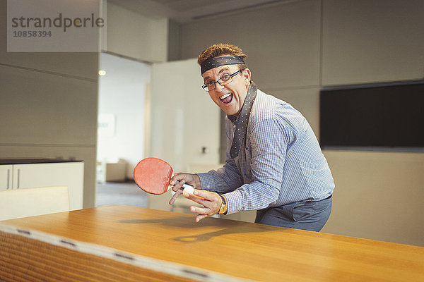 Porträt eines begeisterten Geschäftsmannes  der Tischtennis mit Krawatte um den Kopf im Konferenzraum spielt.