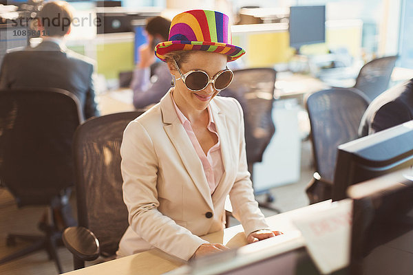 Verspielte Geschäftsfrau mit alberner Sonnenbrille und gestreiftem Hut am Computer im Büro