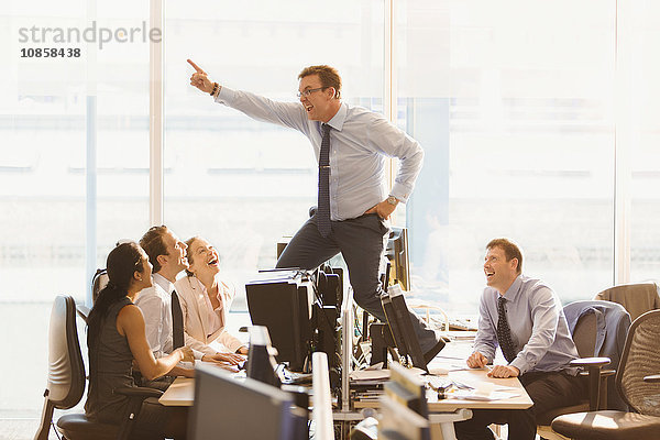 Lachende Kollegen  die übermütige Geschäftsleute auf Schreibtischen im Büro tanzen sehen.
