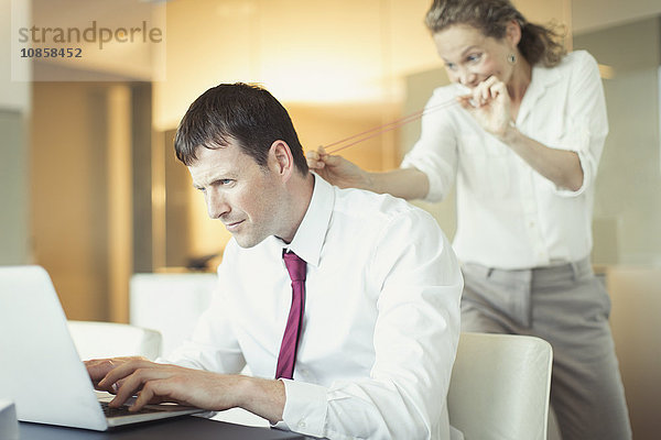 Schelmische Geschäftsfrau zielt mit Gummiband auf ahnungslosen Geschäftsmann am Laptop