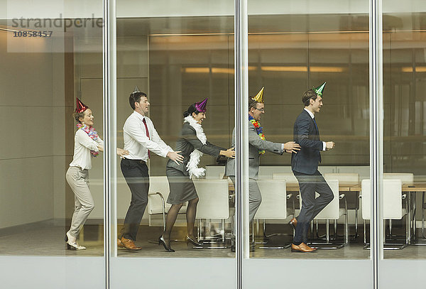 Geschäftsleute mit Partyhüten tanzen in Conga-Linie am Fenster des Konferenzraumes