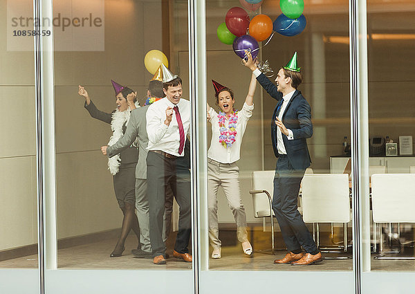 Geschäftsleute mit Partyhüten und Ballontänzen am Fenster des Konferenzraums