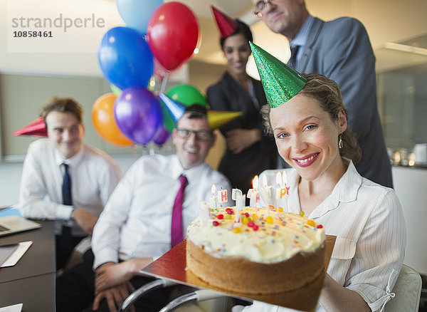 Portrait lächelnde Geschäftsfrau hält Geburtstagskuchen mit Kollegen im Hintergrund