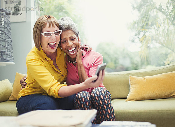 Lachende reife Frauen  die sich umarmen und sich auf dem Sofa niederlassen.