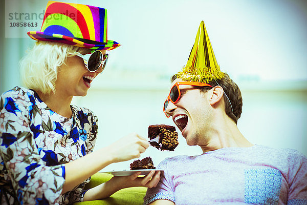 Verspieltes Paar mit Partyhüten und Sonnenbrille beim Schokoladenkuchenessen