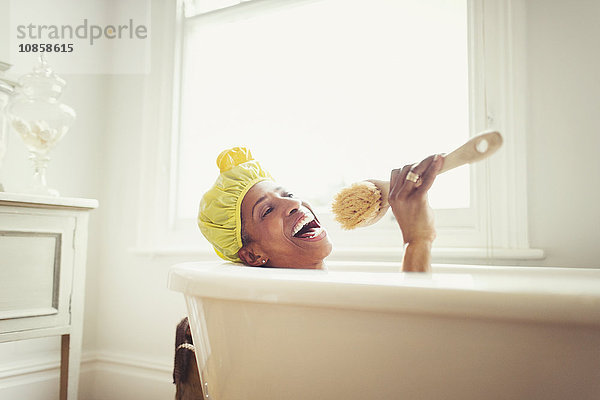 Verspielte reife Frau singt in die Loofah-Bürste in der Badewanne.