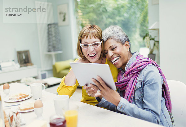 Lachende reife Frauen teilen sich Tablet PCn am Frühstückstisch