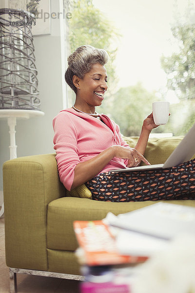 Lächelnde reife Frau mit Laptop und Kaffeetrinken auf dem Sofa