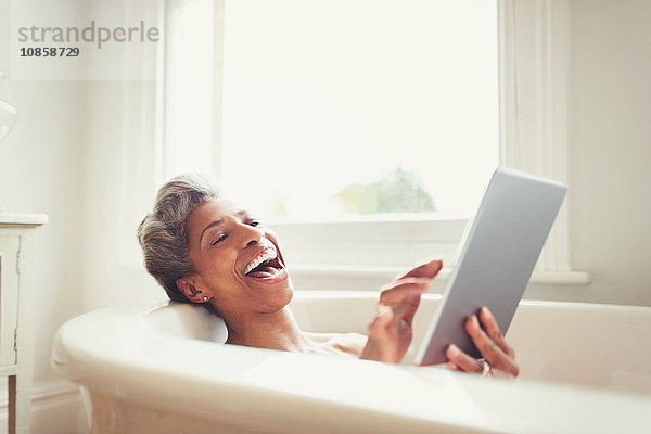 Lachende reife Frau mit digitalem Tablett in der Badewanne