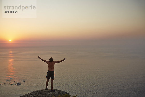Überschwänglicher männlicher Läufer mit ausgestreckten Armen auf Felsen mit Blick auf den Sonnenuntergang im Meer