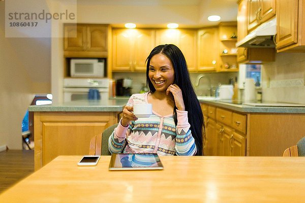 Junge Frau trinkt Kaffee und benutzt digitales Tablett am Küchentisch