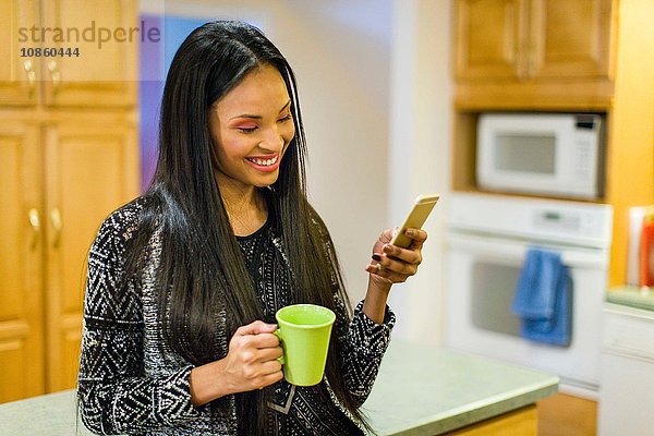 Junge Frau trinkt Kaffee in der Küche und liest dabei Text auf einem Smartphone