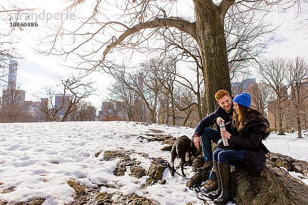 Junges Paar sitzt mit Hund und Trinkflasche im verschneiten Central Park  New York  USA