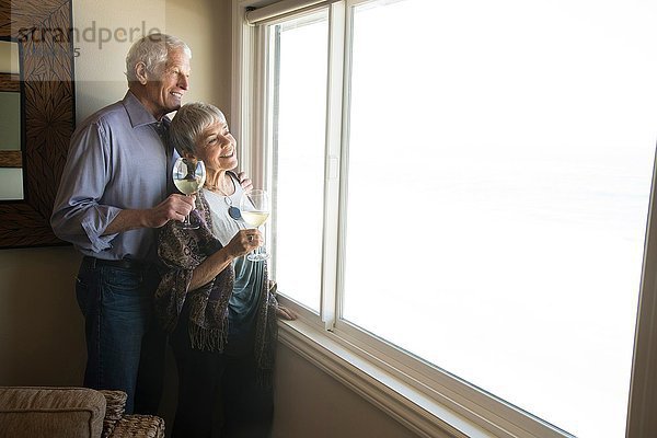 Älteres Ehepaar schaut aus dem Fenster  hält ein Glas Wein in der Hand und lächelt