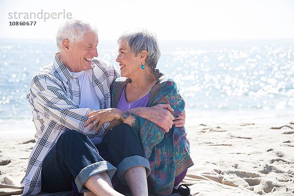 Älteres Paar sitzt am Strand  umarmt sich  von Angesicht zu Angesicht  lächelt