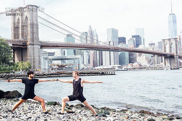 Zwei Männer praktizieren Yoga am Flussufer vor der Brooklyn Bridge  New York  USA