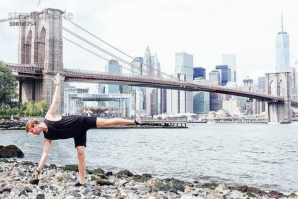 Mittelgroßer erwachsener Mann praktiziert Yoga am Flussufer vor der Brooklyn Bridge  New York  USA