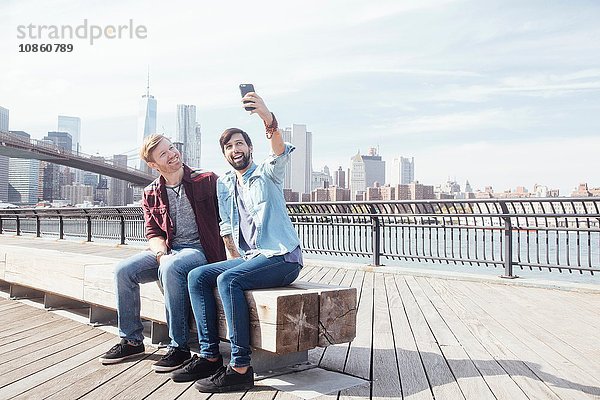 Männliches Paar sitzt am Flussufer bei der Brooklyn Bridge und nimmt Smartphone-Selfie mit  New York  USA