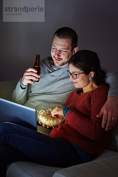 Mittleres erwachsenes Paar sitzt abends auf dem Sofa  isst Popcorn und schaut auf den Laptop