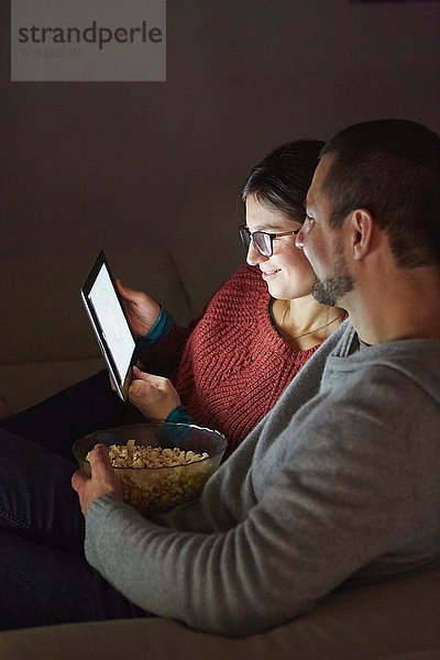 Mittelgroßes erwachsenes Paar sitzt abends auf dem Sofa  isst Popcorn und schaut auf ein digitales Tablet