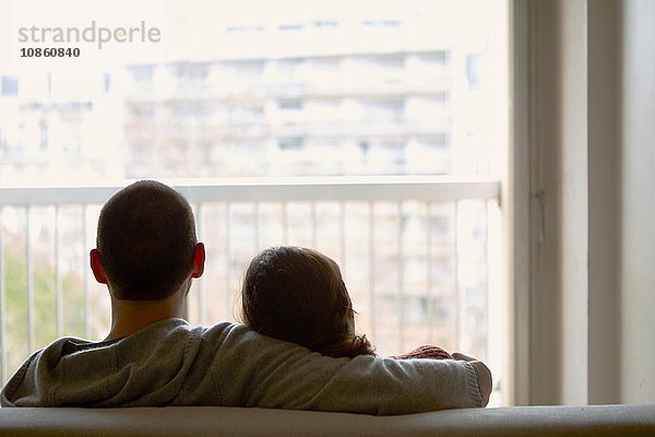 Rückansicht eines auf einem Sofa sitzenden Paares  das aus dem Fenster auf ein Wohnhaus schaut