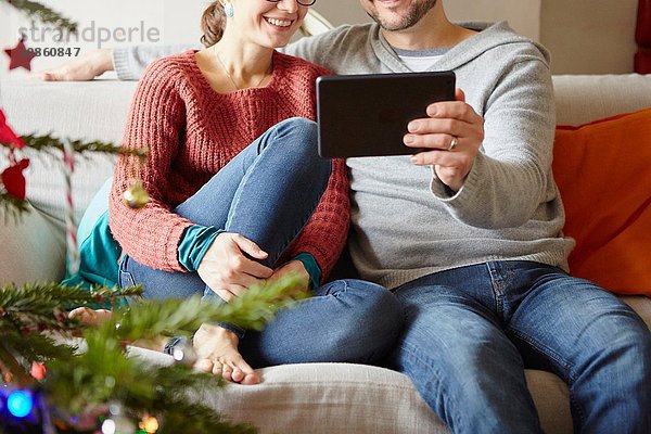 Erwachsenes Paar sitzt zu Weihnachten auf dem Sofa und liest ein digitales Tablet