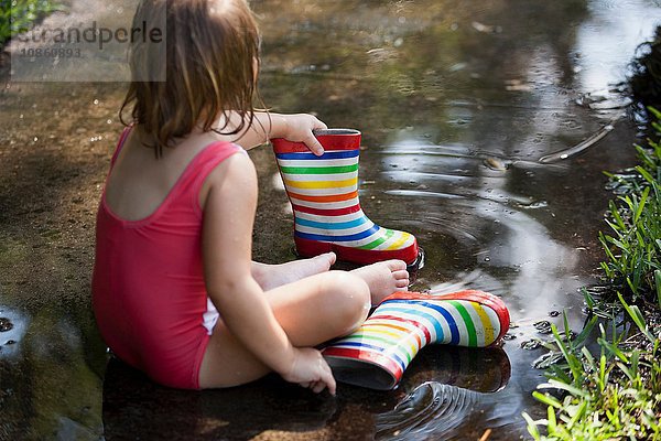 Kind in Gummistiefeln sitzt in Wasserpfütze