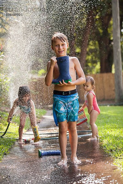 Junge trägt Gummistiefel auf Bürgersteig  Mädchen spielen mit Wasserschlauch im Hintergrund