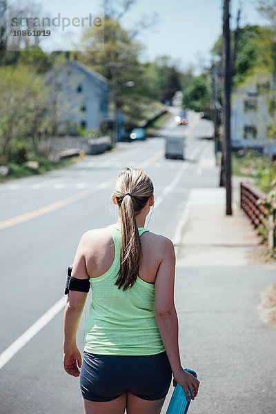 Rückansicht einer Frau  die einen Aktivitäts-Tracker mit einer Wasserflasche trägt und entlang der Straße geht