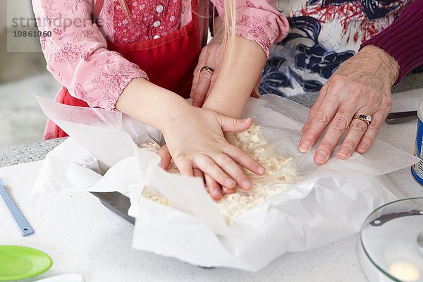 Hände eines Mädchens und einer Großmutter  die Kuchenteig an der Küchentheke verteilen