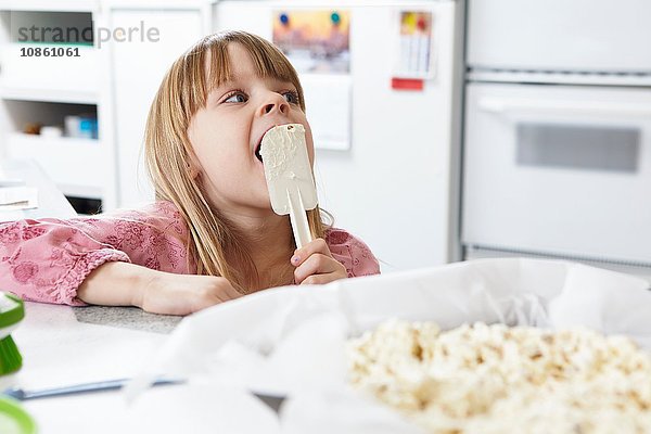 Mädchen leckt Kuchenmasse vom Spachtel an der Küchentheke