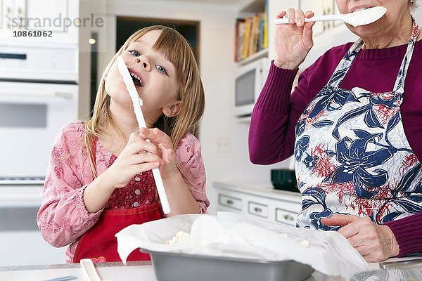 Mädchen und Großmutter lecken Kuchenmischung von Spachteln an der Küchentheke
