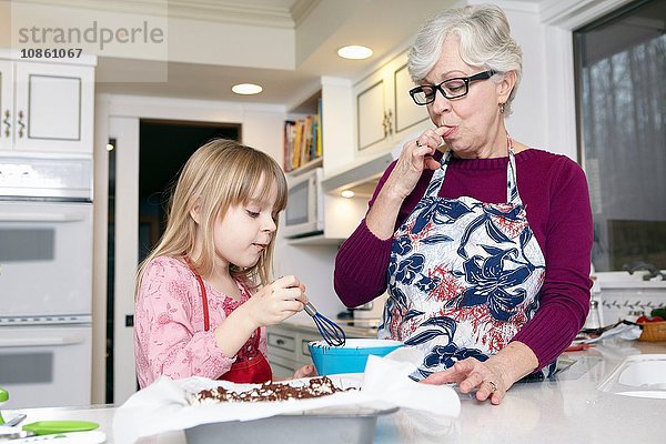Mädchen mischt Kuchen an der Küchentheke  während die Großmutter sich die Finger leckt