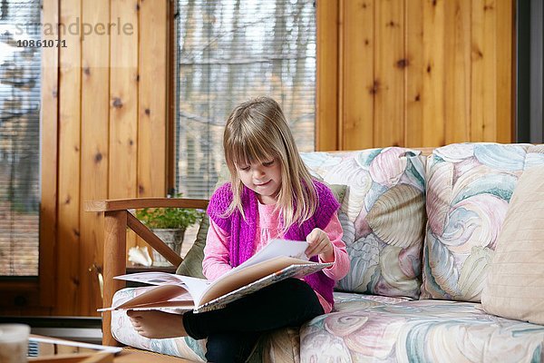 Mädchen sitzt und liest Geschichtenbuch auf Wohnzimmersofa