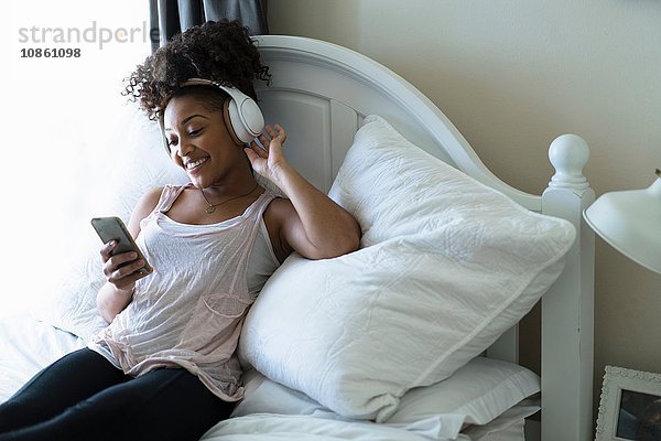 Frau entspannt sich im Bett  trägt Kopfhörer  schaut auf Smartphone