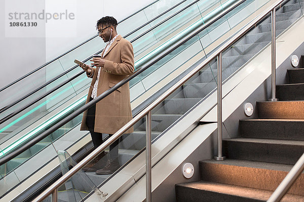 Junger Geschäftsmann fährt die Rolltreppe des Bahnhofs hinunter und liest Smartphone