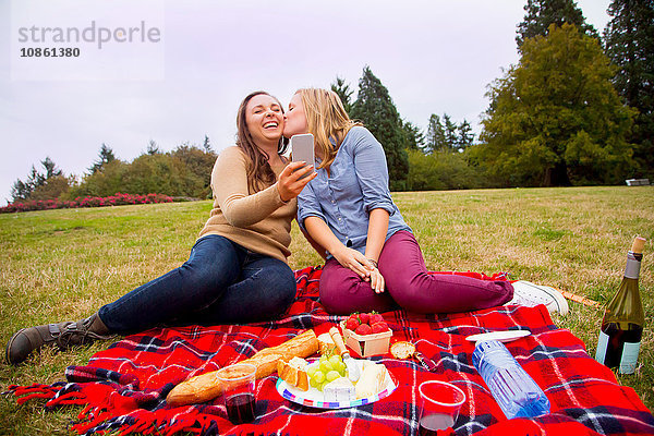 Zwei junge Frauen beim Picknick im Park  beim Herumalbern  beim Selbermachen  mit dem Smartphone