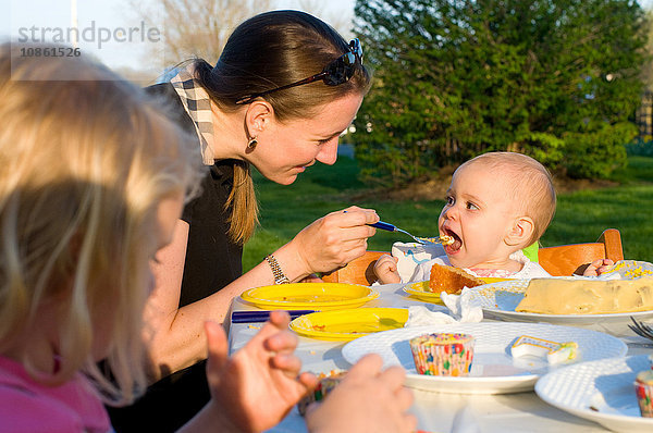 Mutter und Kinder sitzen im Freien  Mutter füttert Baby Mädchen Geburtstagskuchen