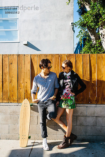 Junges Paar steht zusammen im Freien  Skateboard neben Mann an Wand gelehnt