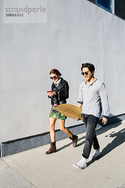 Junges Paar geht gemeinsam im Freien spazieren  Frau benutzt Smartphone  Mann trägt Skateboard